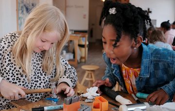 Two children at a bench with jewellery making supplies in front of them. The one child is smiling while using hammer. The other chid is looking to choose an item from the table.