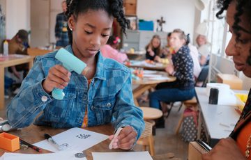 An adult watching a child hold jewellery hammer to stamp a design.