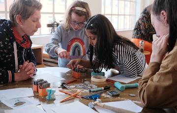 An adult and two children at a table with craft and jewellery making supplies in front of them. The children choosing their supplies.