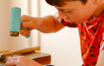 A focused child uses a small hammer to shape a metal piece on a workbench.