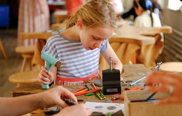 Young child focused on crafting, surrounded by colourful pencils and tools on wooden work bench.