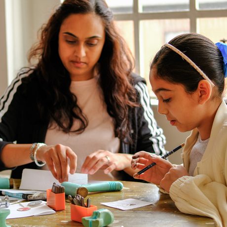 Adult and child engaged in jewellery making activity at a table by a window.