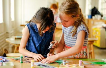 Two children collaborate on a colourful art project using jewellery paint and sticks.