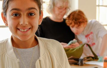 Smiling child shows colourful handmade jewellery pieces, with two people working in the background.