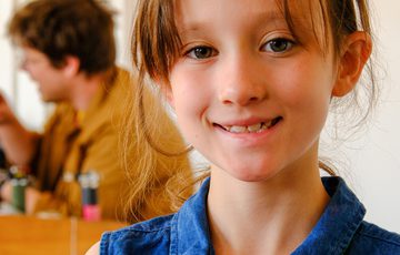 Smiling child holding a colourful necklace pendant, with a blurred person in the background.