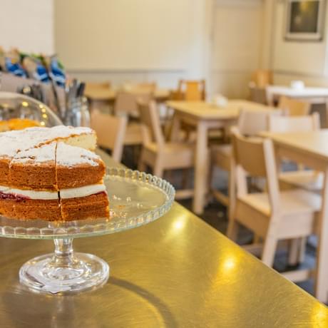 A victoria sponge cake and scones on cake stands on a counter top. In the background are cafe table and chairs.