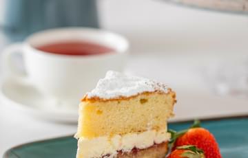 A slice of Victoria sponge cake with strawberries next to it. A cup of tea and teapot is in the background.