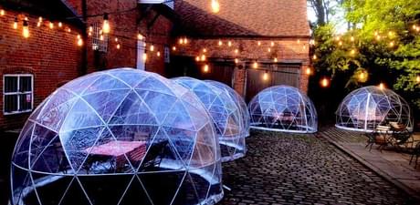 Five igloo pods with tables inside, located in a cobbled courtyard, a brick mill building to the left side, festoon lights hang above.