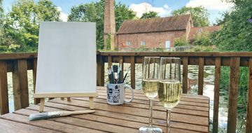 A table with canvas, easel and paint brushes along with two glasses of Prosecco in front of Sarehole Mill and pond.