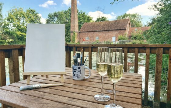 A table with canvas, easel and paint brushes along with two glasses of Prosecco in front of Sarehole Mill and pond.