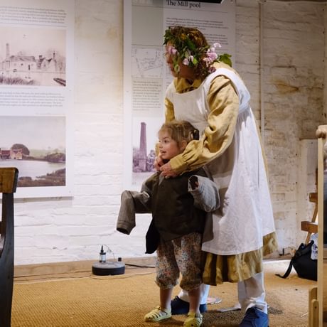 An adult and children inside the mill building wearing historic replica clothing.