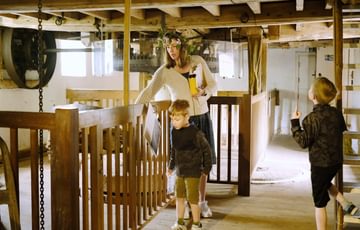 An adult and two children inside the mill with wooden fencing around the machinery.