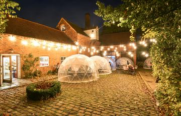 A brick building and cobbled courtyard at night. it is lit up with festoon light. There are igloo pods in the courtyard with seating inside.