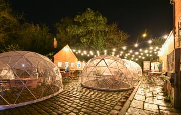 A brick building and cobbled courtyard at night. it is lit up with festoon light. There are igloo pods in the courtyard with seating inside.
