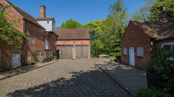 A two storey brick building with single storey outbuilding opposite across a cobbled courtyard.