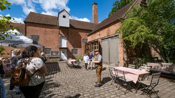 A man is giving a tour of Sarehole Mill to a small group of people. They are standing in the cobbled courtyard.