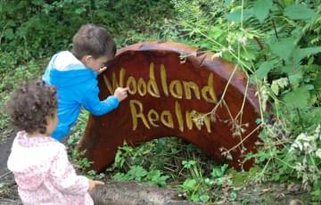 Two children outside, one is pointing to a wooden sign in the ground. The sign reads 'Woodland Realm' and is surrounded by plants.