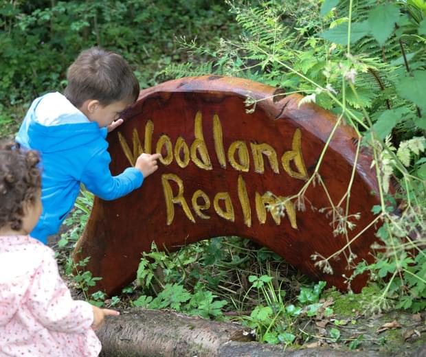 Two children outside, one is pointing to a wooden sign in the ground. The sign reads 'Woodland Realm' and is surrounded by plants.