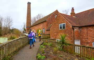 People walking up a path to brick-built water mill building, fence and millpond to the left and planting to the right of the people.