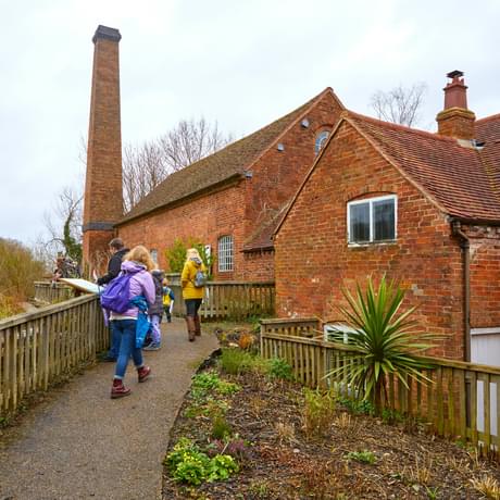 People walking up a path to brick-built water mill building, fence and millpond to the left and planting to the right of the people.