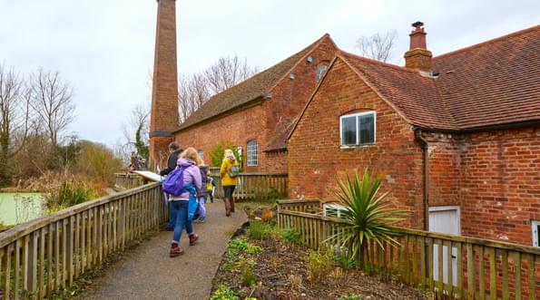 People walking up a path to brick-built water mill building, fence and millpond to the left and planting to the right of the people.