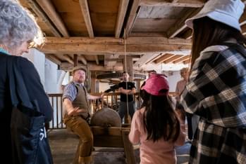 A tour guide is telling a story and the visitors are standing listening to him. They are inside the mill and above them are the wooden beams and boards that make up the ceiling.