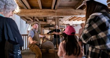 A tour guide is telling a story and the visitors are standing listening to him. They are inside the mill and above them are the wooden beams and boards that make up the ceiling.