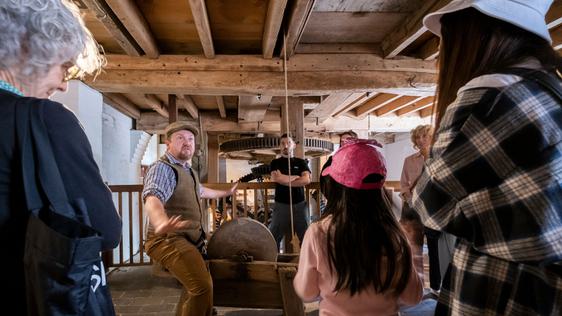 A tour guide is telling a story and the visitors are standing listening to him. They are inside the mill and above them are the wooden beams and boards that make up the ceiling.