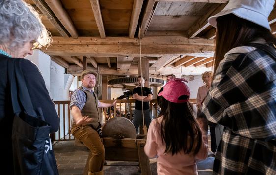 A tour guide is telling a story and the visitors are standing listening to him. They are inside the mill and above them are the wooden beams and boards that make up the ceiling.