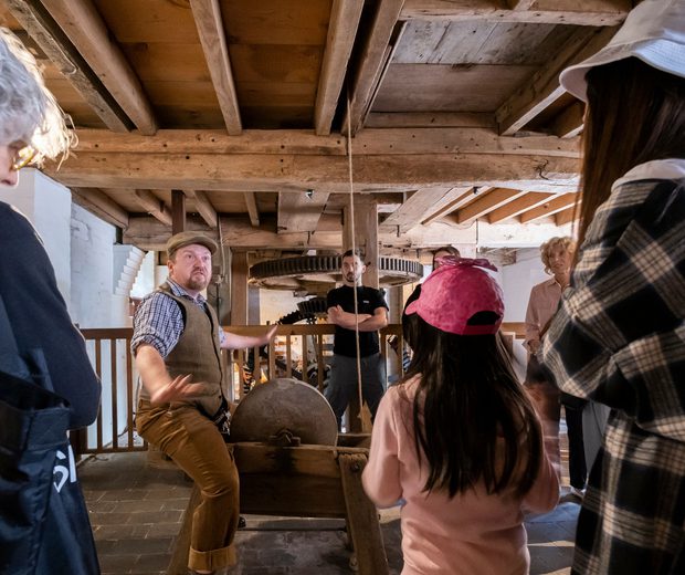 A tour guide is telling a story and the visitors are standing listening to him. They are inside the mill and above them are the wooden beams and boards that make up the ceiling.