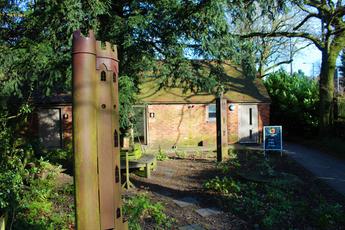 Two sculptures of tall thin towers in the gardens surrounded by tress and bushes, a small building is in the background