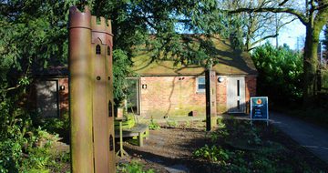 Two sculptures of tall thin towers in the gardens surrounded by tress and bushes, a small building is in the background