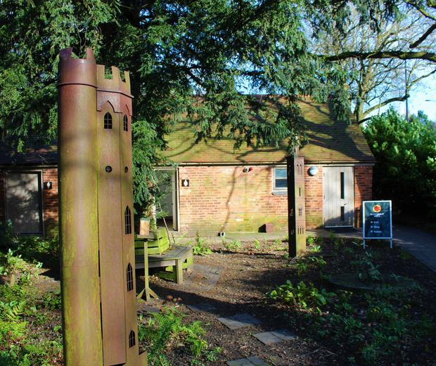 Two sculptures of tall thin towers in the gardens surrounded by tress and bushes, a small building is in the background