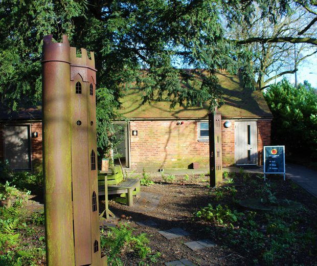 Two sculptures of tall thin towers in the gardens surrounded by tress and bushes, a small building is in the background
