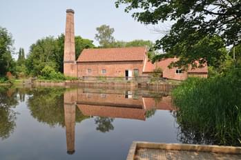 A mill building with chinmey behind a pond which has its reflection in the water.