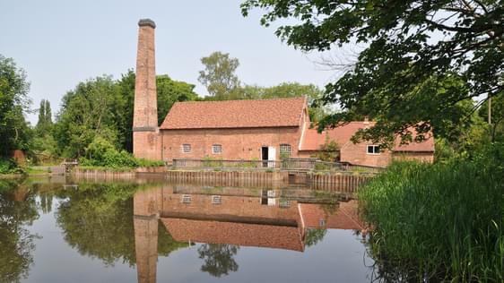 A mill building with chinmey behind a pond which has its reflection in the water.