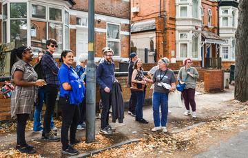 A group of people standing at the side of a road, with houses behind. They are smiling and looking at something out of view.