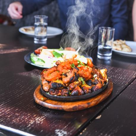 Plate of steaming hot food on a wooden table, with a person in the background.