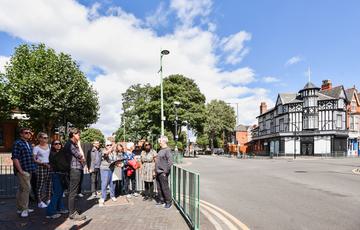 A group of people standing at a road junction, a tour guide is pointing at something out of view. There are buildings and trees behind the group.
