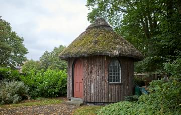 A small round building in garden with thatched room and arched door and window.