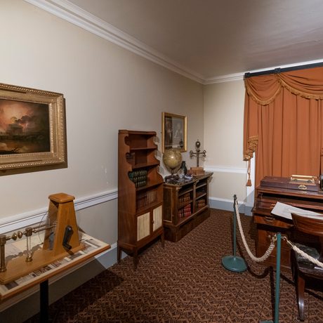 A study with wooden desk, sideboard and furniture, a painting hangs of the wall and the draped curtains are closed.
