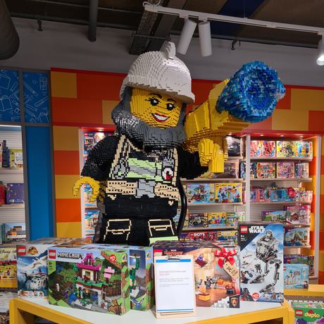 A large figure made out of lego is standing on a dispaly unit with many different boxes of lego for sale.
