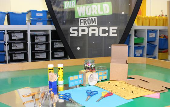 Craft items such as pens, paper, scissors and glue on a table with an example crafted satellite. In the background a banner reads 'Our World From Space'.