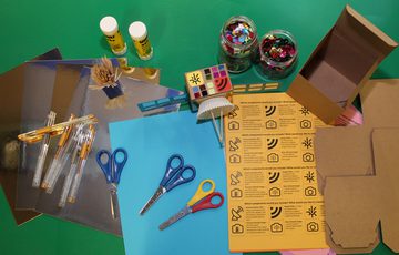 Craft items such as pens, paper, scissors and glue on a table with an example paper based satellite.
