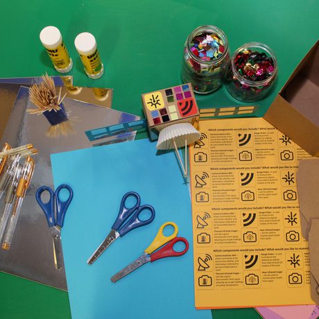 Craft items such as pens, paper, scissors and glue on a table with an example paper based satellite.