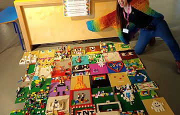 A large number of LEGO baseplates with lLEGO designs laid out on a floor. A staff member in a colourful cardigan is next to them with their arms out.