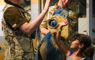 A girl high-fives a puppet dinosaur operated by two people in safari outfits.