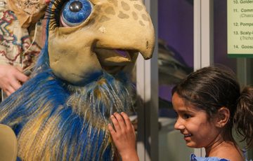A young girl joyfully touches a large, colourful dinosaur puppet with blue feathers.