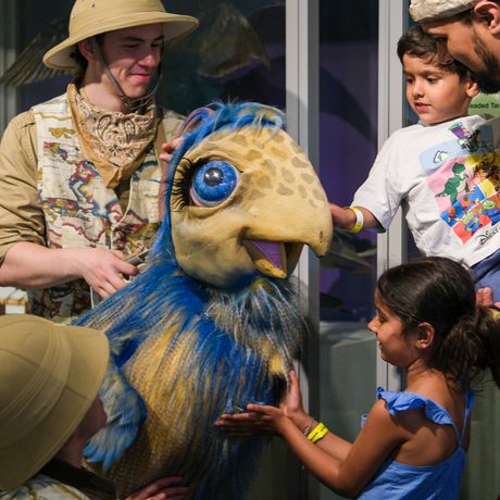 Children interact with a large, colourful dinosaur puppet operated by a performer in safari attire.
