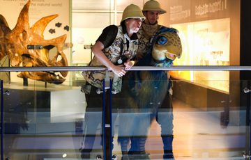 Two explorers in safari hats interact with a dinosaur puppet near a fossil exhibit.